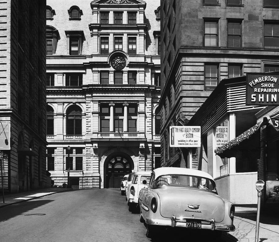 Black-and-white photo of a city street lined with parked 1950s cars, historic buildings, and storefronts, including a shoe repair shop and an employment booth, with a large ornate building in the background.
