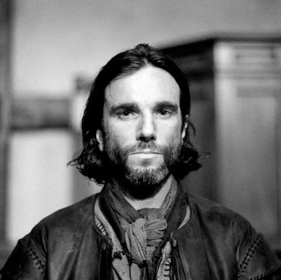 Black-and-white portrait of a man with shoulder-length hair and a beard, wearing a scarf and a leather jacket, looking directly at the camera with a neutral expression.