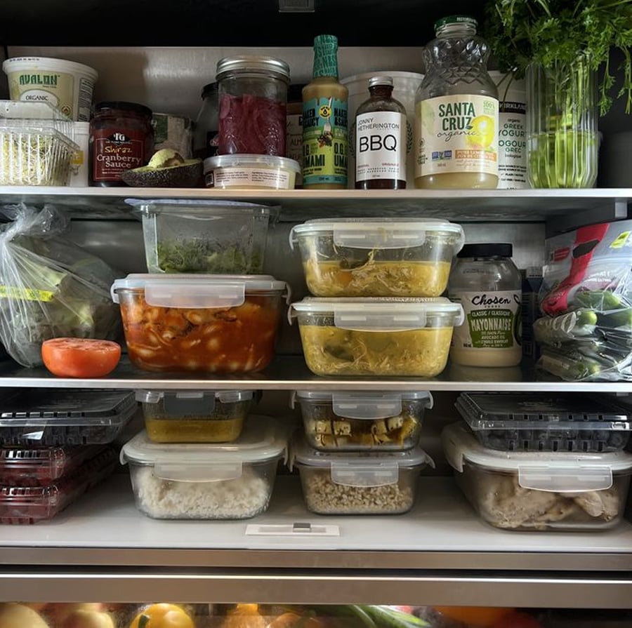 A neatly organized refrigerator filled with clear containers of prepared food, fresh vegetables, berries, condiments, and drinks. Parsley and bottles are on the top shelf; fruits and vegetables are in drawers at the bottom.