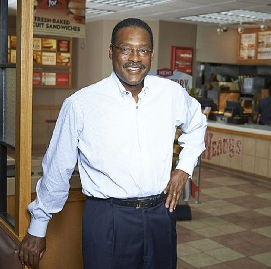 A man wearing glasses, a light blue dress shirt, and dark pants stands smiling inside a Wendy’s restaurant. He rests one hand on a wooden divider, with a counter and menu boards visible in the background.