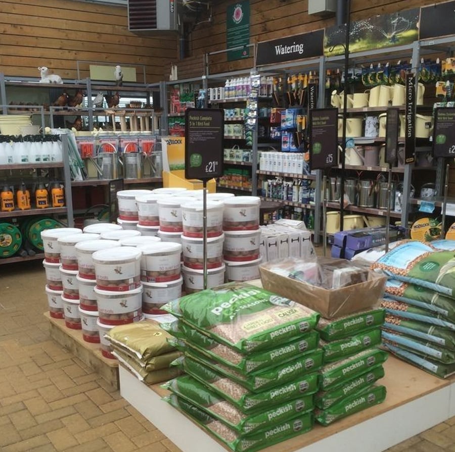A garden center display shows stacks of green bags of pet-friendly organic pebbles, white tubs, and shelves with various watering cans, plant care products, and gardening supplies. Signs display prices above the products.