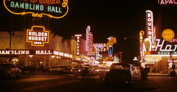 A lively nighttime street scene in Las Vegas, featuring glowing neon signs for casinos and hotels such as Golden Nugget and Hotel Apache, with cars and people visible under the bright lights.