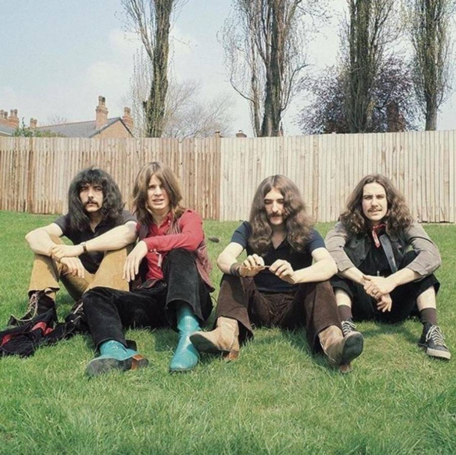 Four men with long hair sit on grass in front of a wooden fence and leafless trees. They wear 1970s-style clothing, including boots and jackets, and look relaxed while facing the camera.