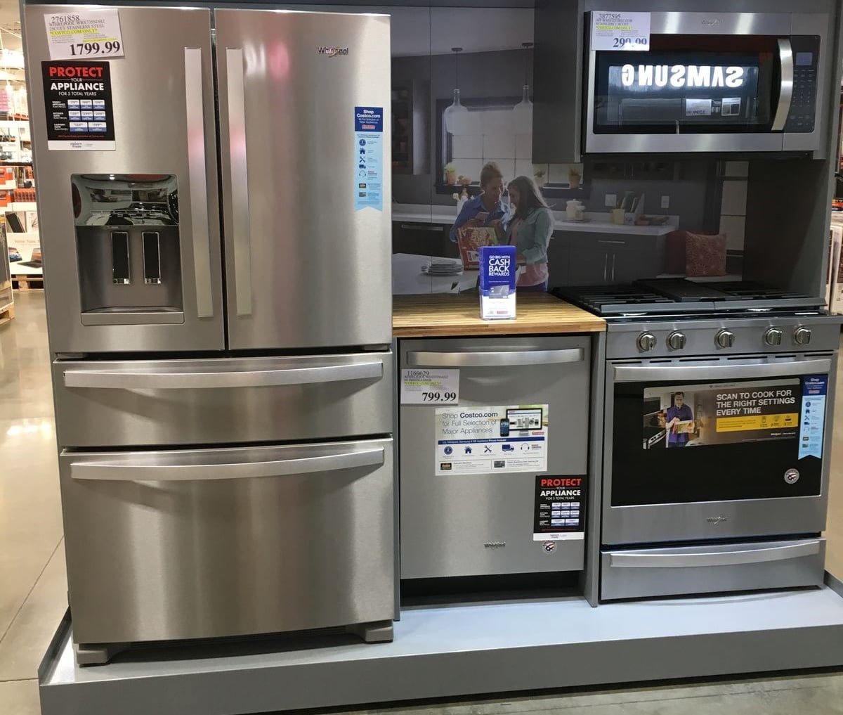 A display of stainless steel kitchen appliances including a French door refrigerator, a dishwasher, a freestanding oven with stove, and a microwave, set up in a store with price tags visible.