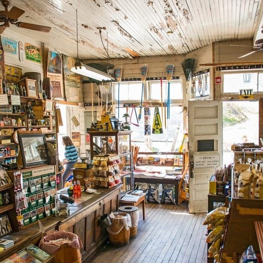 A cluttered, rustic general store interior with wooden floors, shelves stocked with goods, snacks, and vintage signs on the walls. Sunlight streams in through large front windows, highlighting the nostalgic, cozy atmosphere.