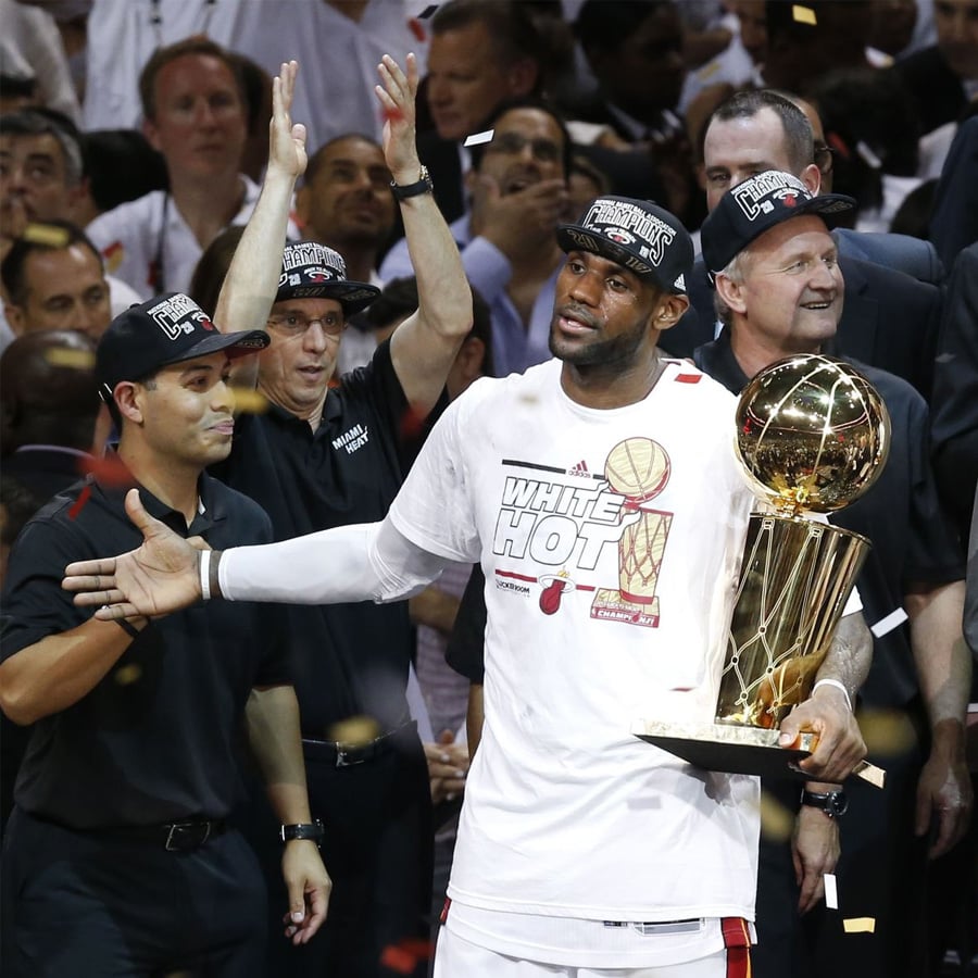 A basketball player in a "White Hot" shirt and championship cap holds the NBA Finals trophy amid celebrating teammates and staff, surrounded by confetti on a crowded court.