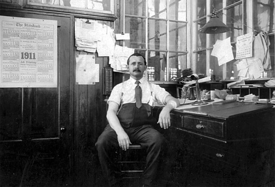 A man with a mustache sits in an old-fashioned office, wearing suspenders and a tie. Papers and calendars cover the walls, and a large desk with documents and a lamp is beside him. The calendar shows the year 1911.