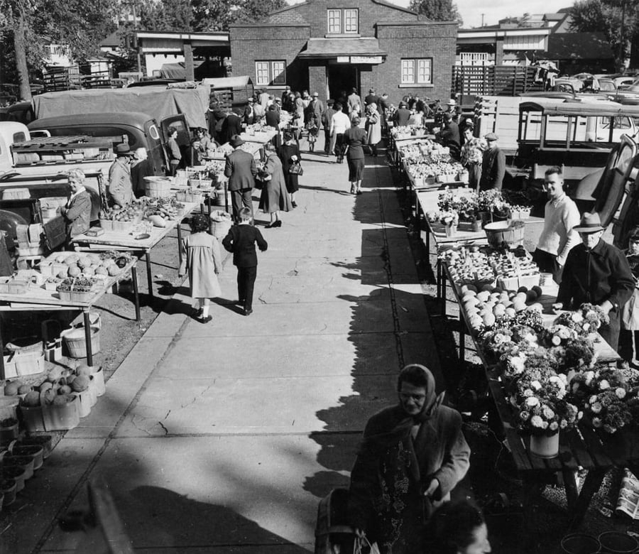 Black-and-white photo of a busy outdoor market with people shopping for produce and flowers at vendor stalls on both sides of a wide walkway, with a brick building visible in the background.