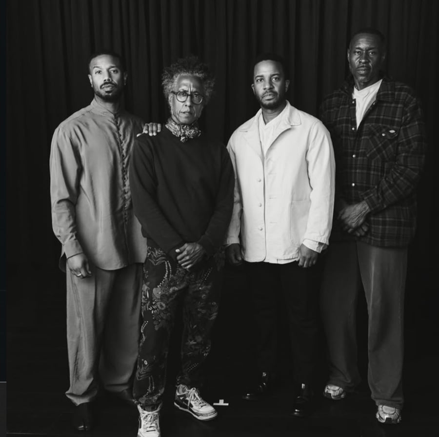 Four men stand side by side in front of a dark curtain. They wear varied styles of clothing, from casual to semi-formal. The image is in black and white, and the men face the camera with serious expressions.