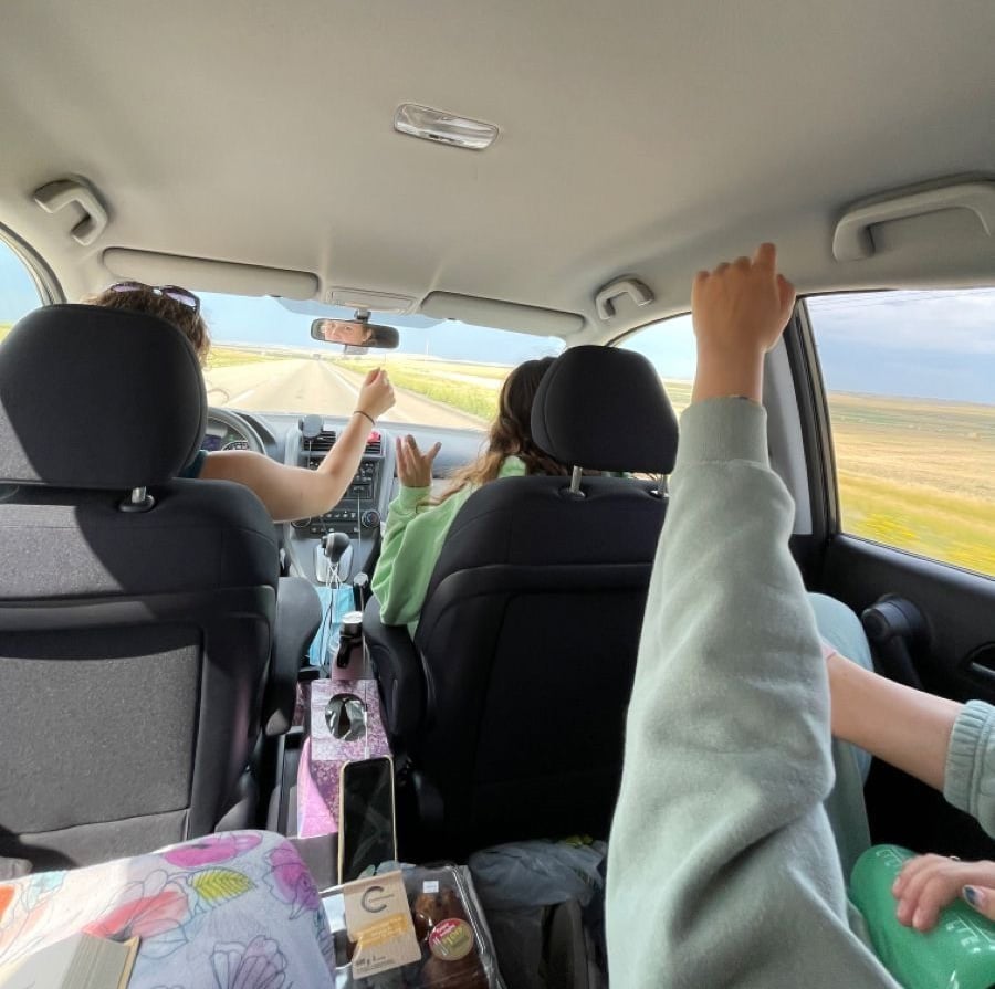 Three people are on a road trip in a car, seen from the backseat. Two are raising their arms joyfully, while the driver has one hand on the wheel. The road and fields stretch out ahead under a partly cloudy sky.