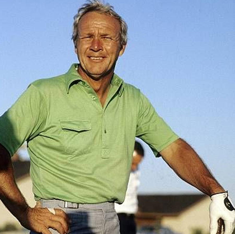A smiling older man in a green short-sleeve polo shirt and gray pants stands outdoors on a sunny day, wearing a golf glove and holding a golf club. The sky is clear and blue, and houses are visible in the background.