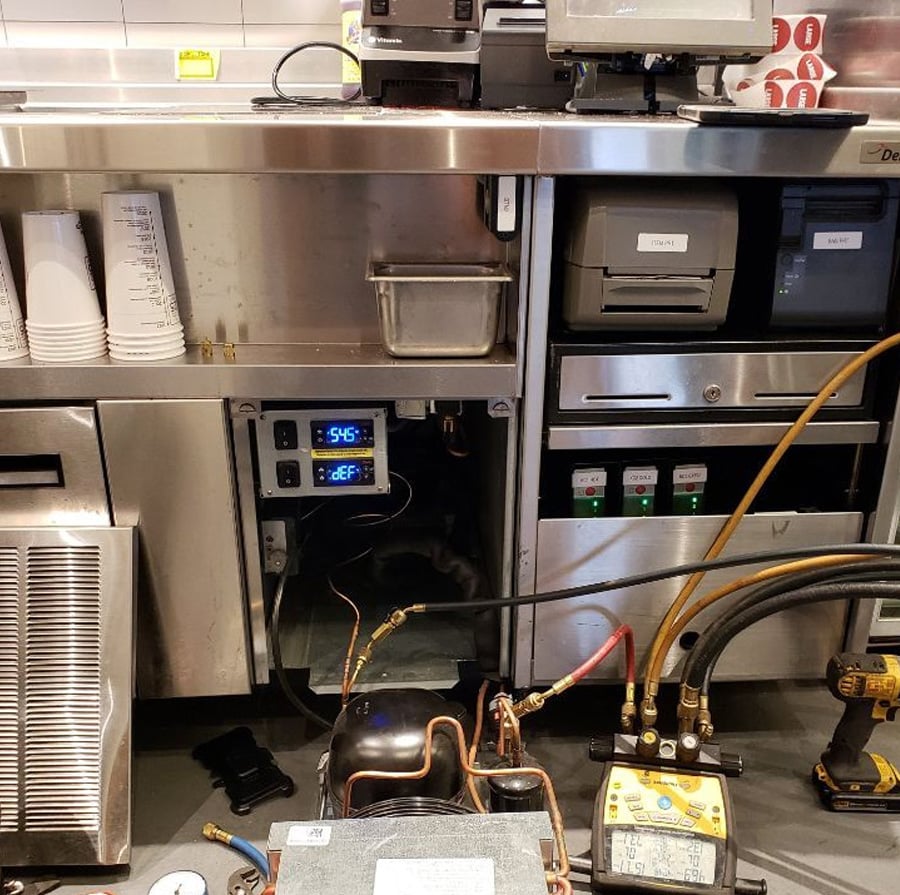 Commercial kitchen counter with exposed appliances and repair tools, including gauges and hoses. An open panel reveals electrical components and digital displays showing temperatures. Paper cups and a receipt printer are on the counter.