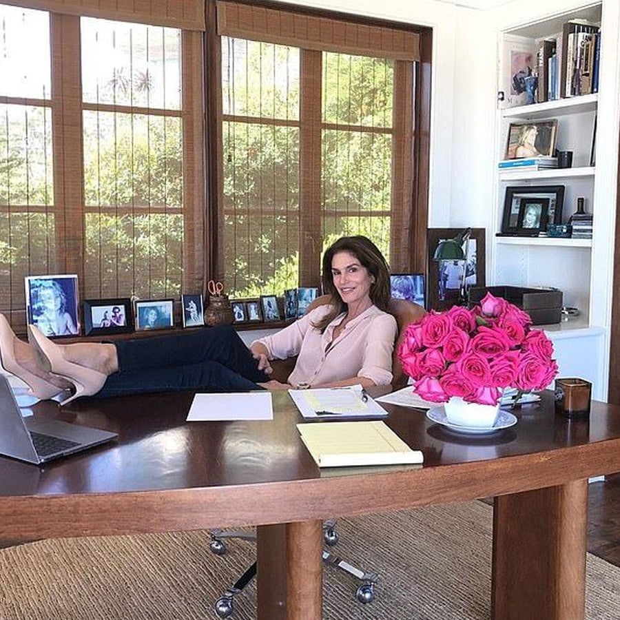 A woman sits at a wooden desk with her feet up, smiling. The desk has papers, a laptop, a notepad, and a vase of pink roses. Behind her are large windows, family photos, shelves, and framed pictures.