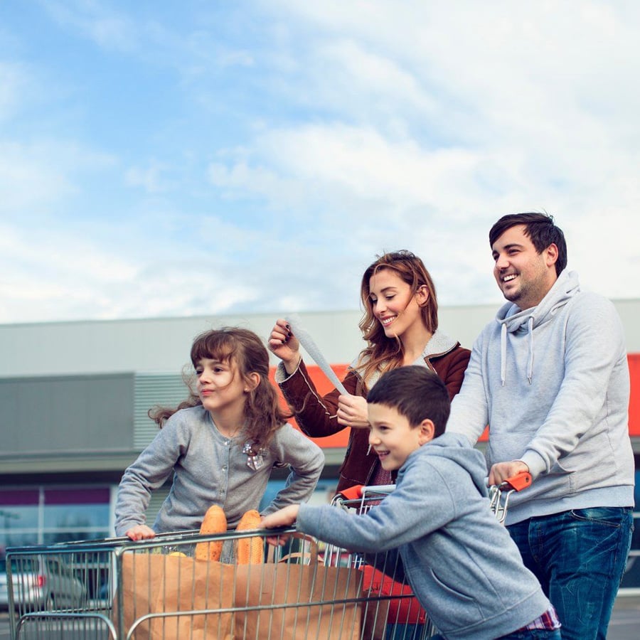 A smiling family with two young children stands outside a store, pushing a shopping cart filled with groceries and baguettes. The parents and children look happy and are dressed in casual clothes.