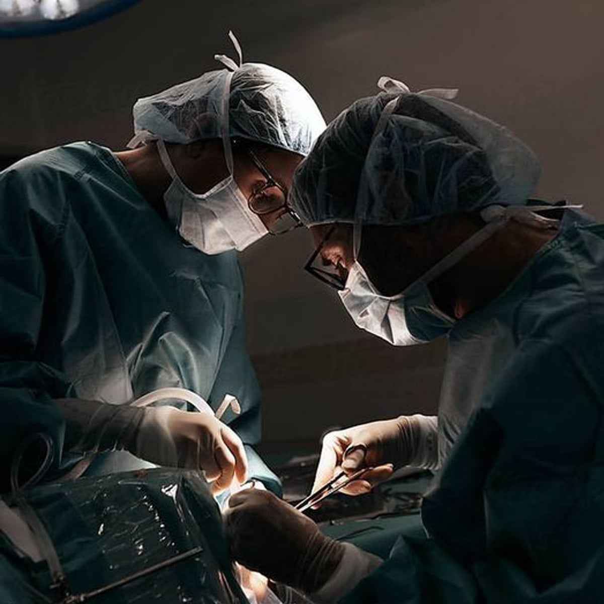 Two surgeons wearing scrubs, masks, gloves, and hair covers perform surgery under bright lights, focusing intently on the procedure with surgical instruments. The background is dimly lit.