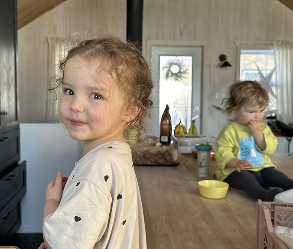 A young girl with curly hair and a beige shirt with black hearts smiles at the camera in a kitchen, while another child in a green shirt sits on the counter eating. The kitchen is bright and cozy.