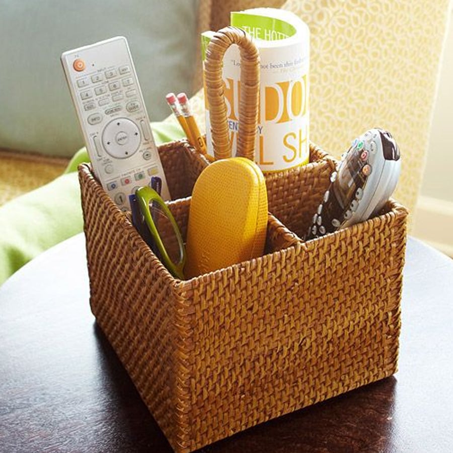 A woven basket organizer sits on a table, holding a TV remote, a phone, scissors, a yellow glasses case, pencils, and a magazine. The basket has separate compartments for each item.