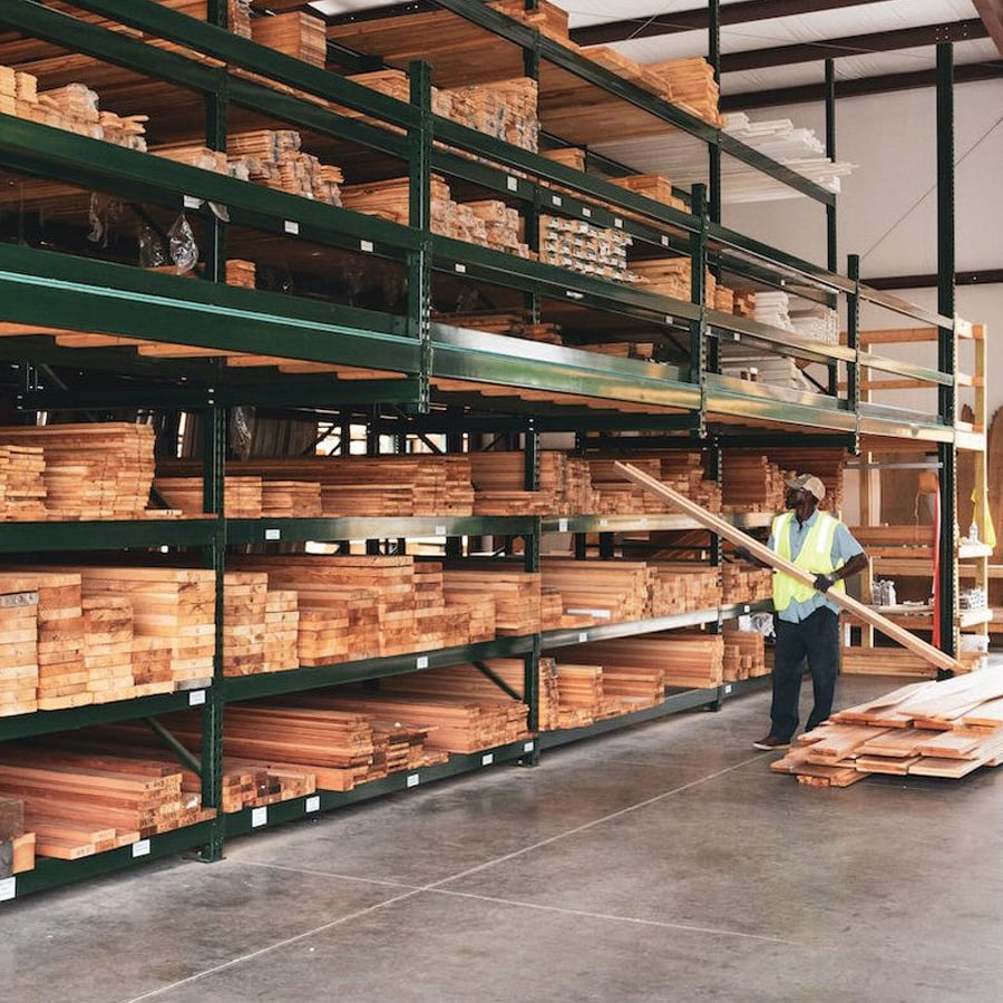 A worker in a yellow safety vest moves a long wooden board in a warehouse filled with neatly stacked lumber on green metal shelves.