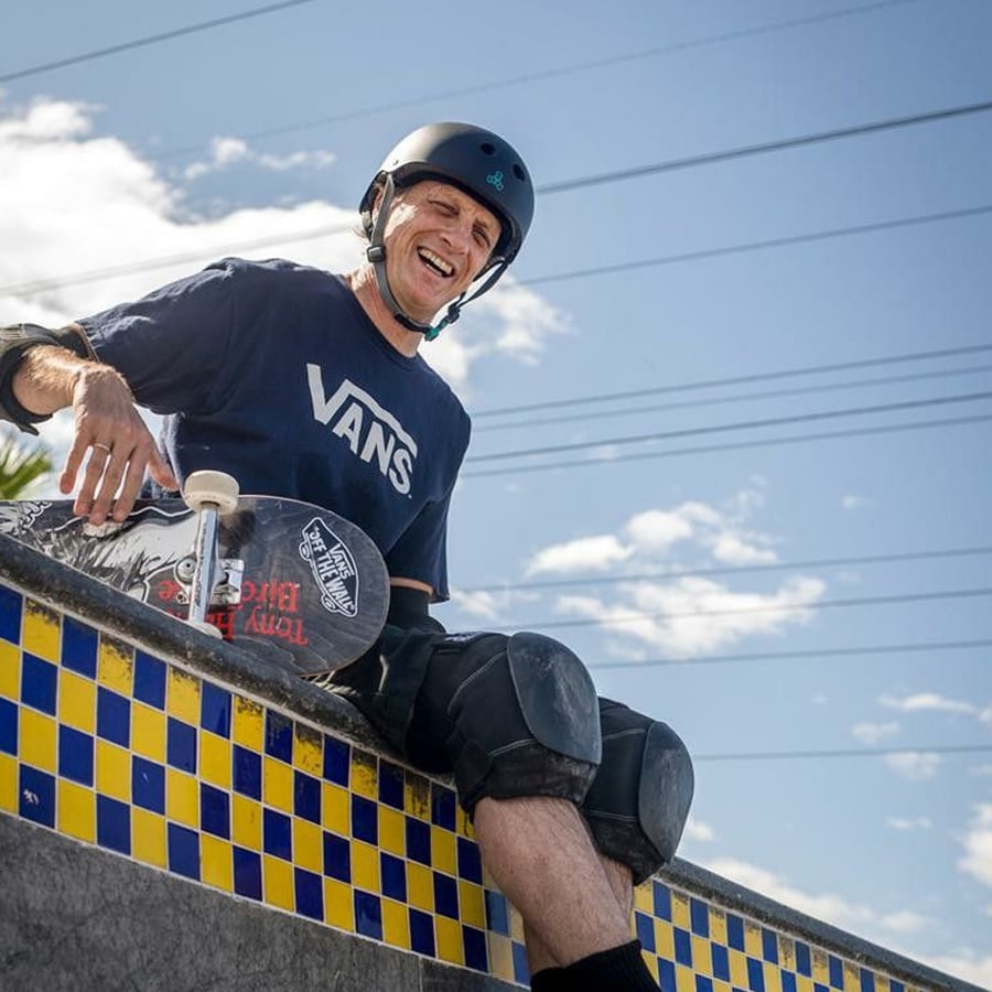 A man wearing a helmet, kneepads, and a Vans t-shirt sits on the edge of a skatepark ramp, smiling, with a skateboard beside him. The sky is blue with scattered clouds and power lines in the background.