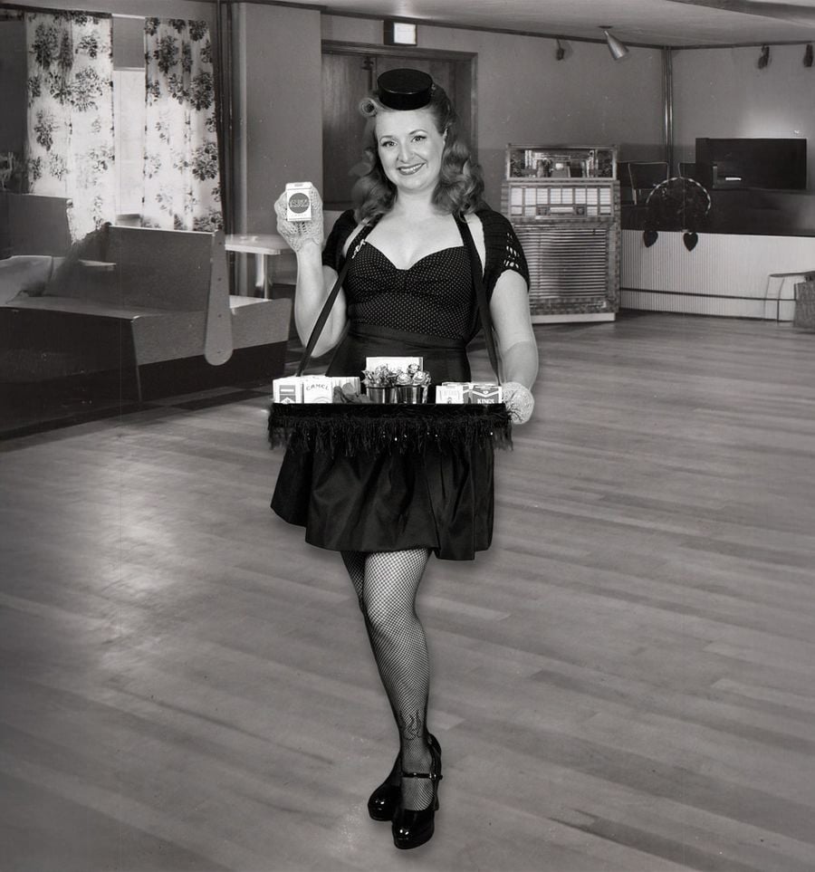 A woman in vintage attire smiles while holding up a pack of cigarettes. She wears a tray around her neck stocked with candy, cigarettes, and gum, standing in a retro-styled room with wood floors and a jukebox in the background.