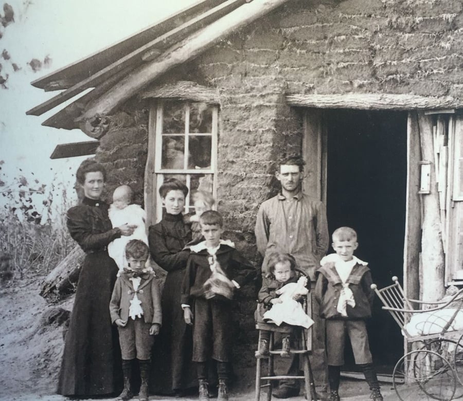 A black-and-white photo of a family with four children and a baby standing outside a rustic sod house. The adults and children wear old-fashioned clothing, and a small stroller is visible next to the group.