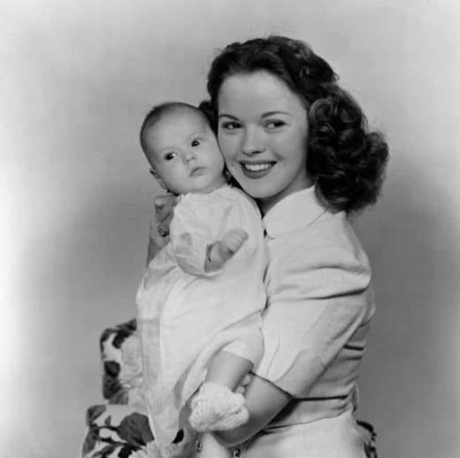 A woman with curly hair, wearing a light-colored dress, holds a baby dressed in a white outfit. Both are looking slightly off camera and smiling. The background is plain and the photo is in black and white.
