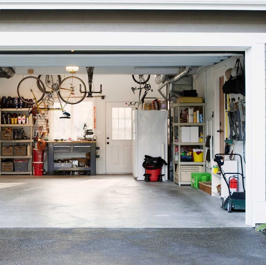 A clean, organized garage with shelves holding tools, boxes, and supplies. Bicycles hang from the ceiling, a refrigerator is against the back wall, and a lawn mower and vacuum are near the open garage door.