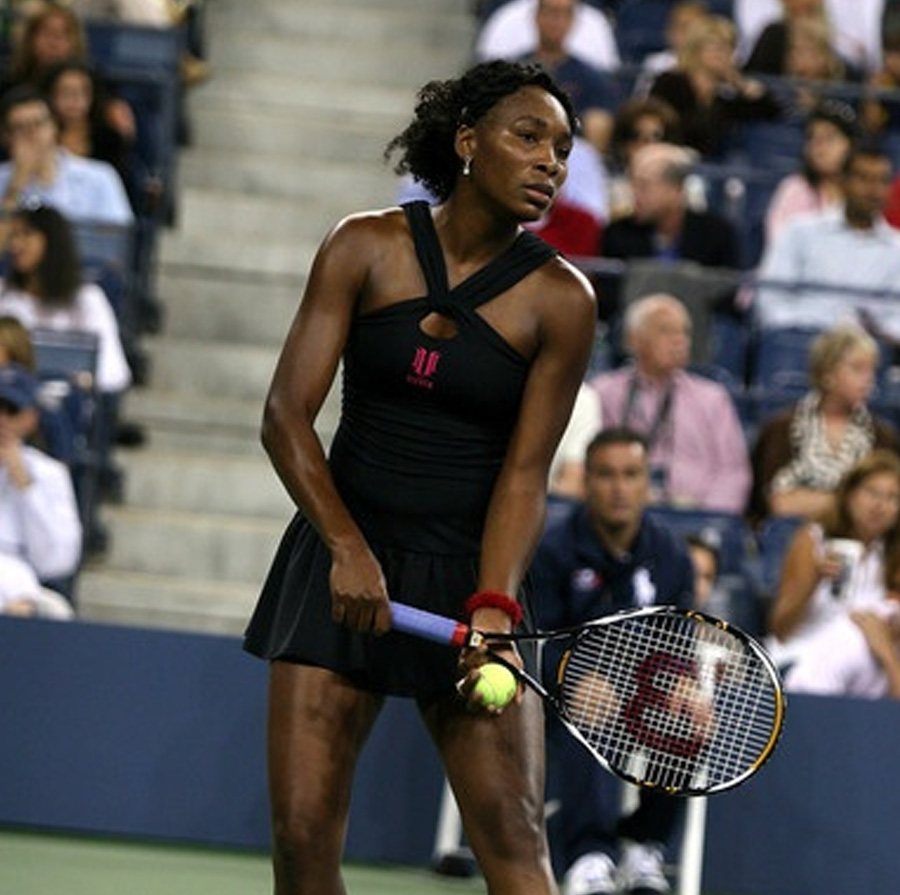 A tennis player in a black dress prepares to serve on an outdoor court, holding a tennis ball and racket, with spectators watching from the stands in the background.