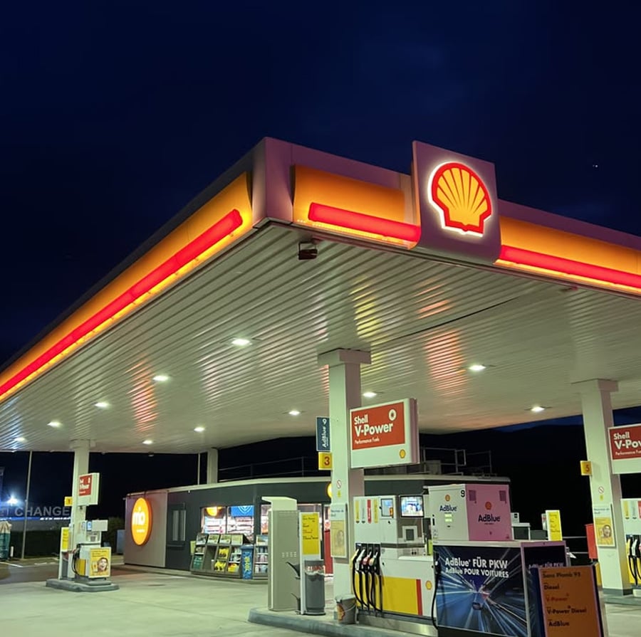 A Shell gas station is brightly lit at night, with fuel pumps visible under the canopy and the illuminated Shell logo on display. Signs for Shell V-Power and other fuel options are also visible.