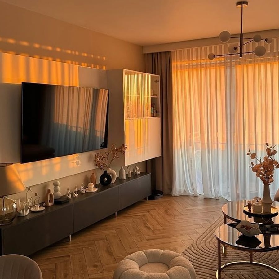 Modern living room with warm golden sunlight streaming through sheer curtains, casting soft shadows. A TV is mounted above a sleek cabinet, with decorative objects and vases on display. Cozy, inviting atmosphere.