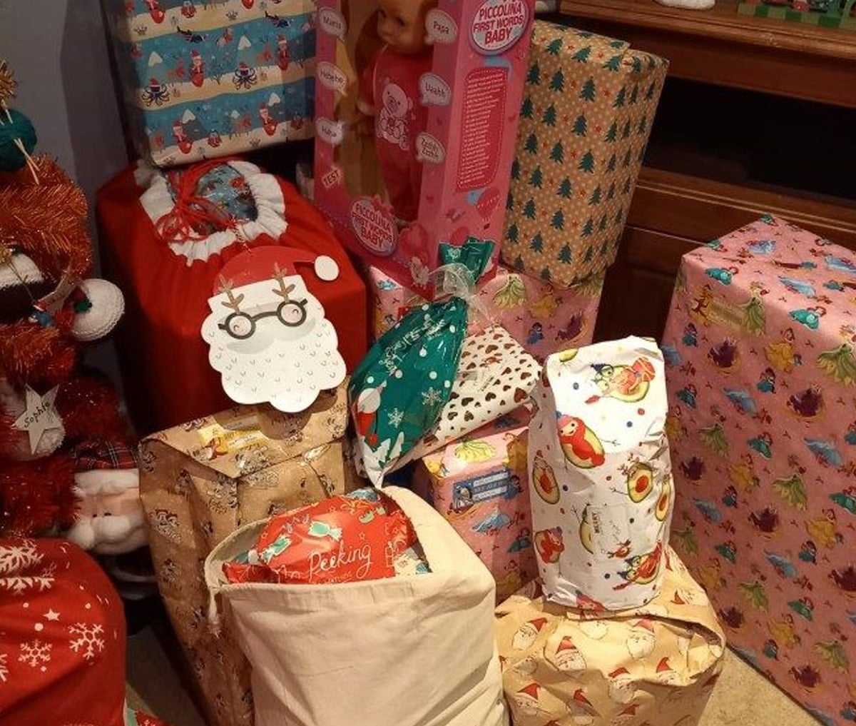 A collection of wrapped Christmas presents in various colorful papers, including one with a Santa face tag, a visible doll toy in a pink box, and festive decorations surrounding the gifts.