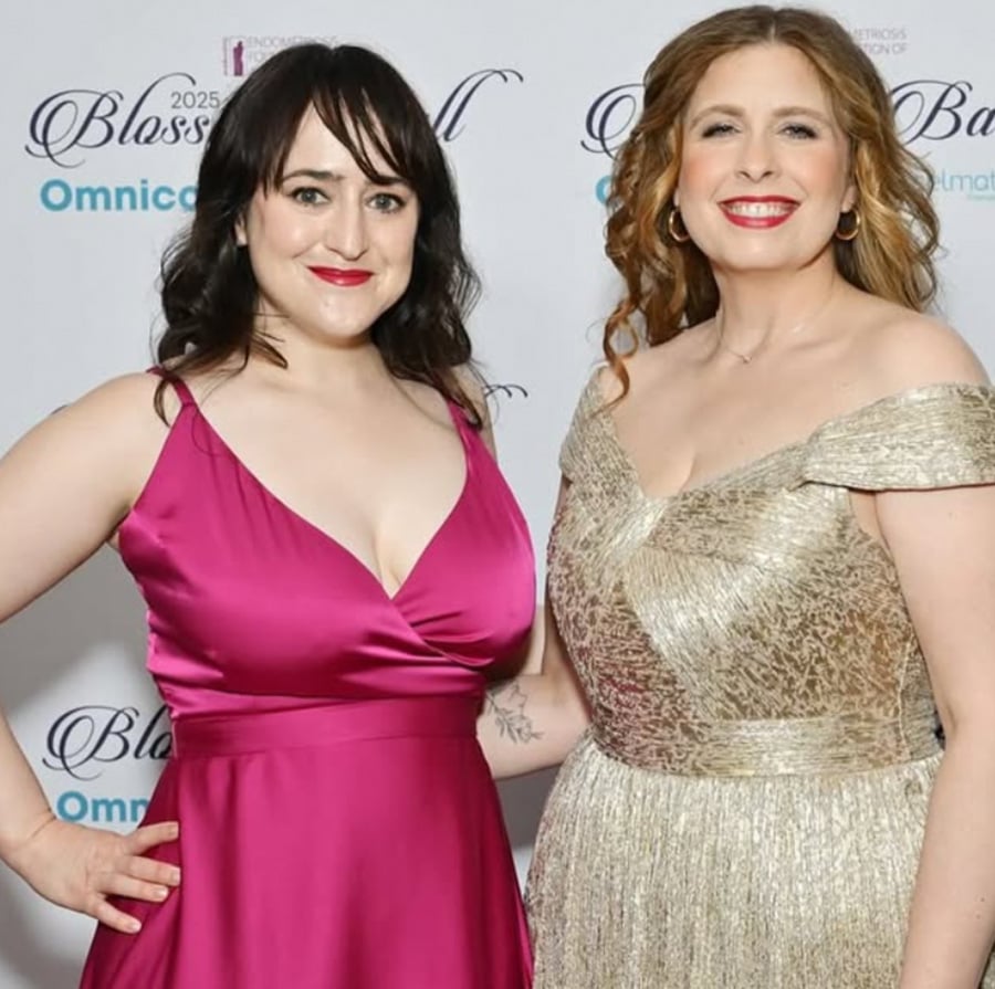 Two women in formal dresses smiling at a gala event. One wears a sleeveless magenta dress, the other a gold off-shoulder gown. Event signage with "Blossom Ball" is visible in the background.