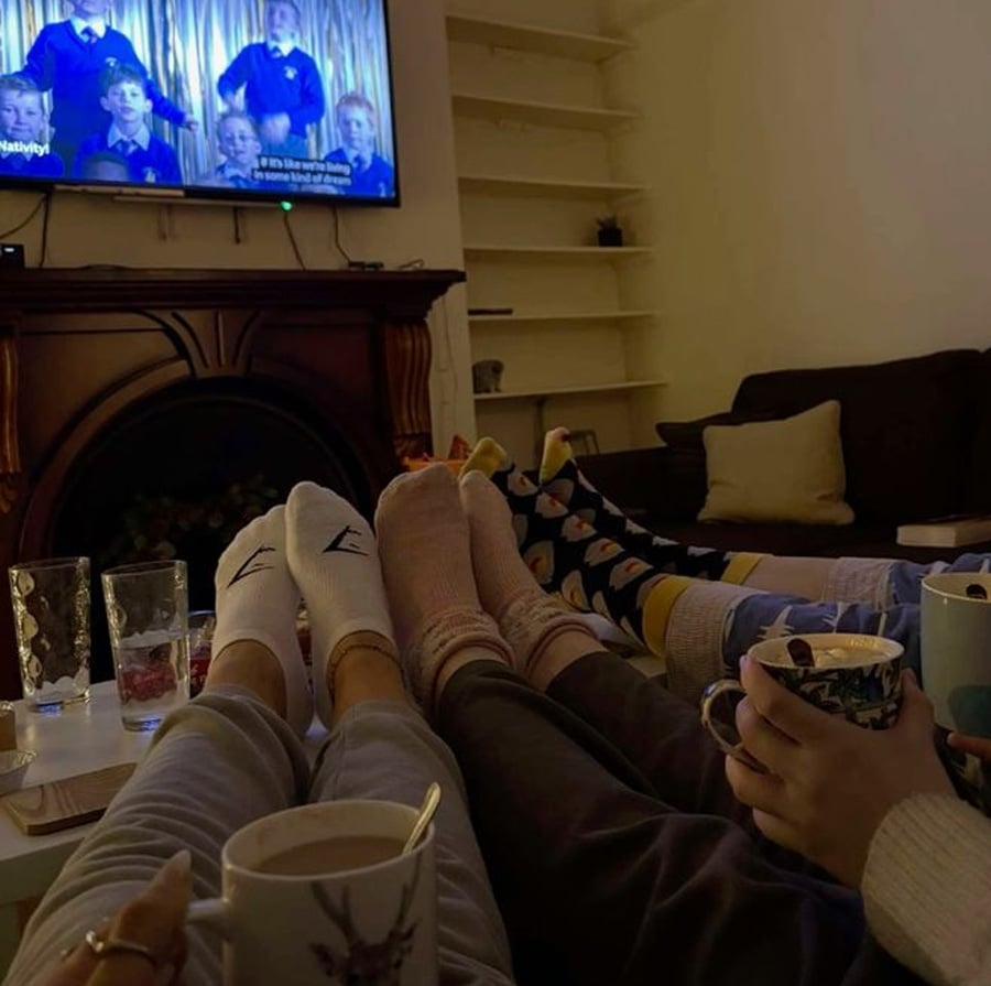 Four people with colorful socks relax on a sofa with hot drinks, their feet up toward a coffee table. A TV plays a show in the background, and the room has cozy lighting with some shelves and cushions visible.