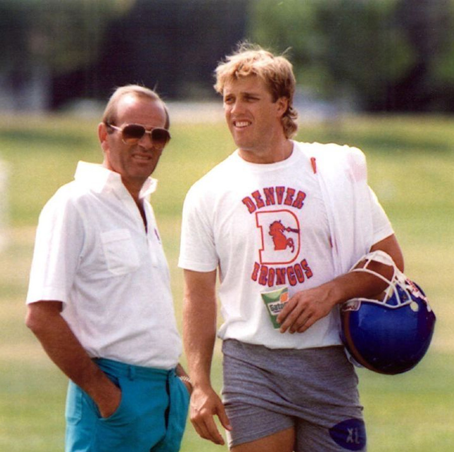 Two men stand outdoors on a grassy field. One wears sunglasses, a white shirt, and teal pants. The other wears a "Denver Broncos" t-shirt, gray shorts, and holds a blue football helmet. Both are smiling and talking.