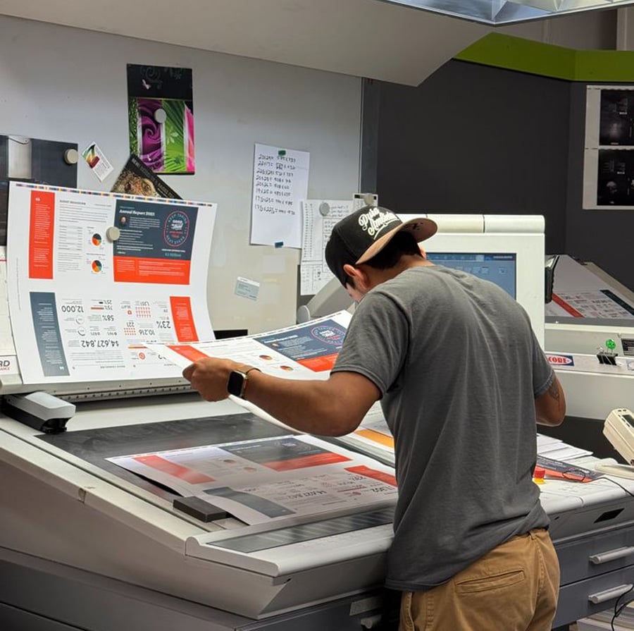 A person wearing a cap and gray t-shirt inspects printed materials at a large, modern printing station, surrounded by colorful printouts and monitors in a print shop.