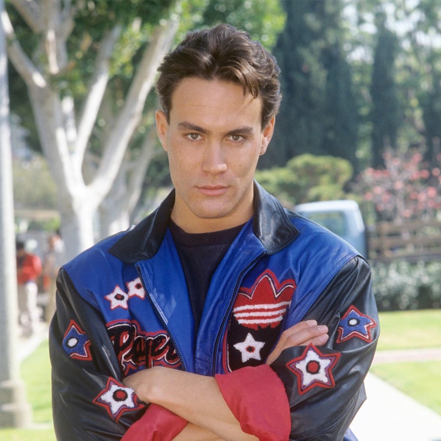 A man with short dark hair stands outdoors with arms crossed, wearing a blue and black jacket decorated with red, white, and black stars and a heart. Trees and greenery are visible in the background on a sunny day.
