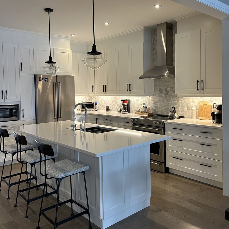 Modern kitchen with white cabinets, stainless steel appliances, a central island with sink and seating for three, pendant lights above, and light gray tile backsplash. Countertops are white and decor is minimal.