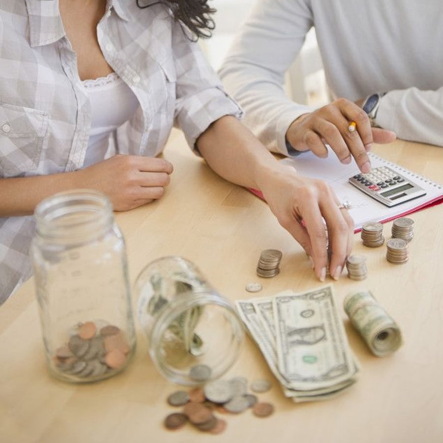 Two people sit at a table counting money, with jars of coins, dollar bills, and a calculator on the surface. One person writes in a notebook while the other arranges coins.