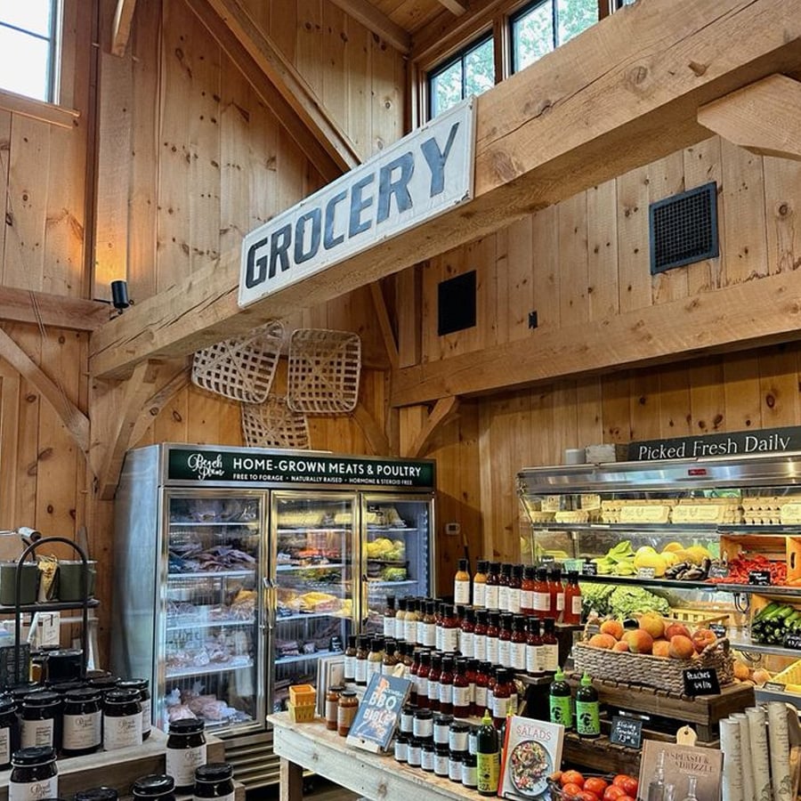 A rustic grocery store interior with wooden beams, a "GROCERY" sign, shelves of fresh produce, bottled goods, and a refrigerator stocked with meats and poultry. Natural light enters through high windows.