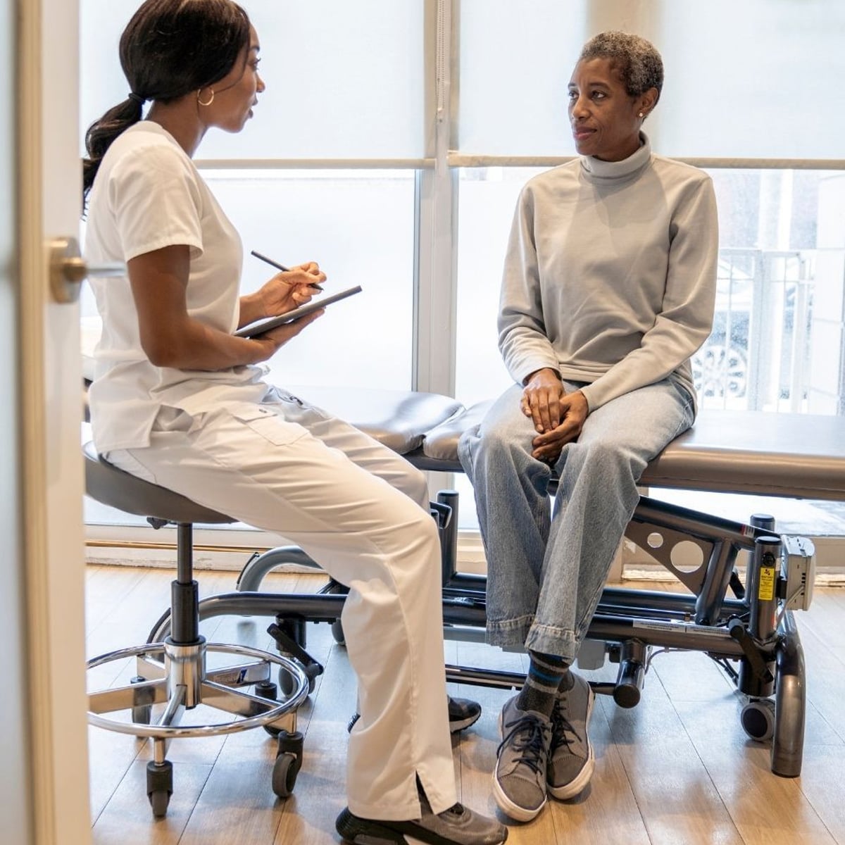 A healthcare professional in white scrubs sits with a clipboard, talking to a patient sitting on an exam table in a medical office with large windows.