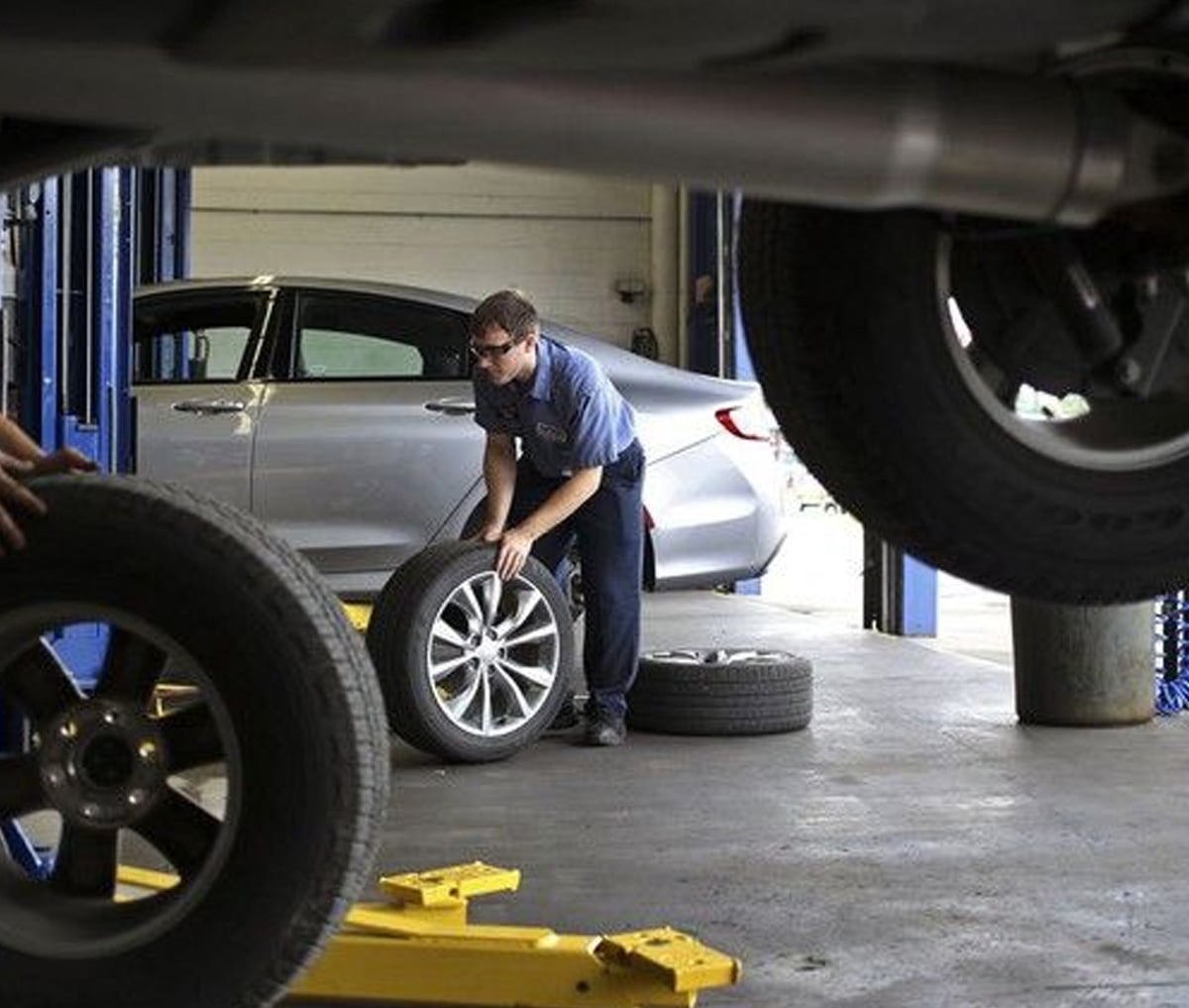 A mechanic in blue work clothes lifts a tire near a silver sedan inside an auto repair shop. Other car tires and a vehicle on a lift are visible in the foreground.