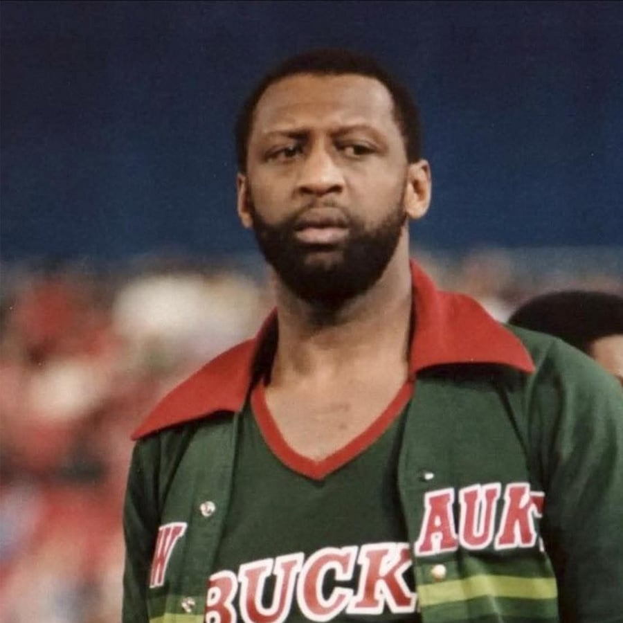 A basketball player wearing a green Milwaukee Bucks warm-up jacket with red accents stands on a court, looking focused. The background is blurred with spectators visible.