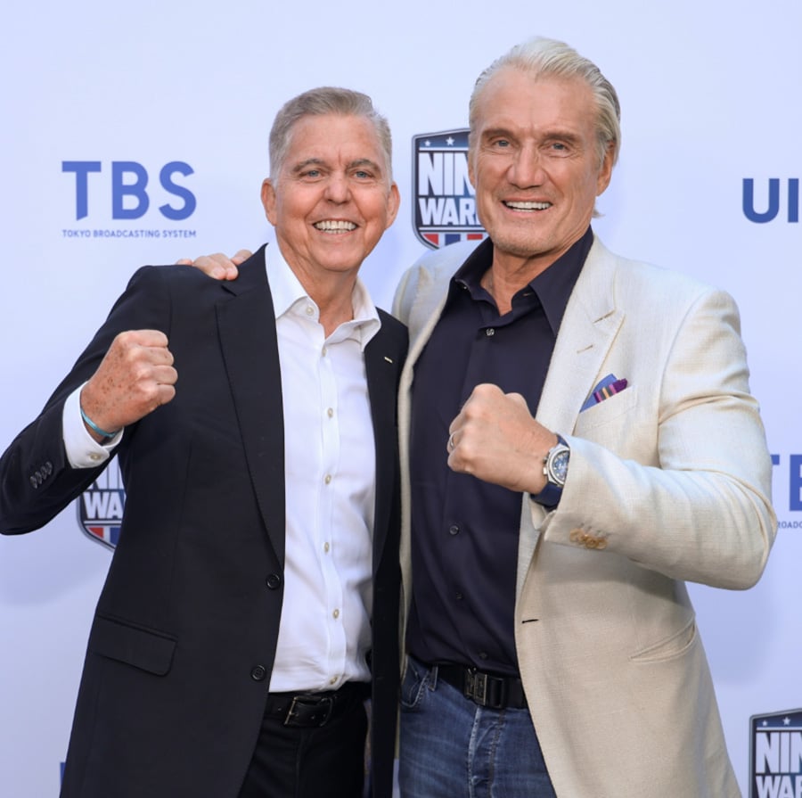 Two men in suits stand smiling with their arms around each other, both holding up fists in a celebratory pose, in front of a backdrop with logos including TBS and Ninja Warrior.