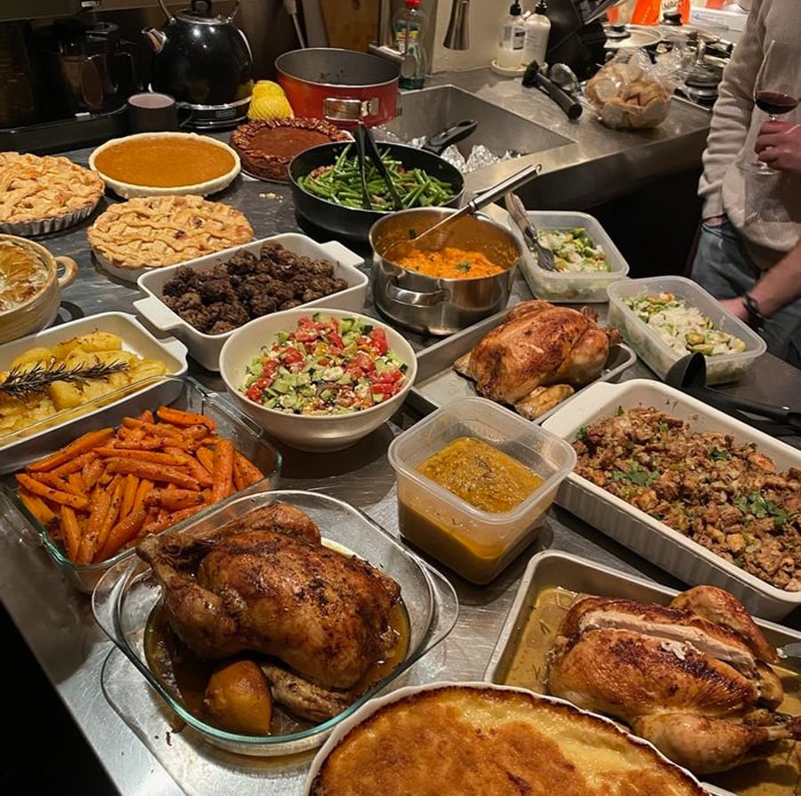 A kitchen counter filled with various homemade dishes, including roasted chickens, pies, roasted carrots, salads, stuffing, green beans, and other side dishes, suggesting a holiday or festive meal.