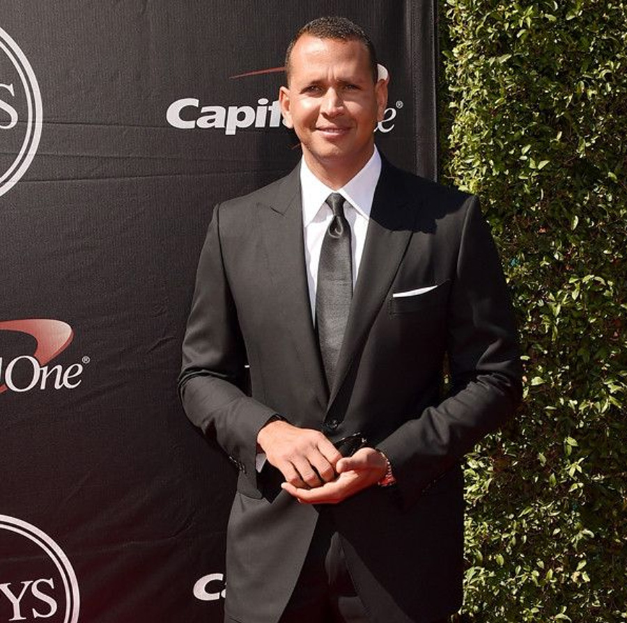 A man in a black suit, white shirt, and gray tie stands on a red carpet in front of a black backdrop with logos and greenery. He is smiling and posing for the camera.