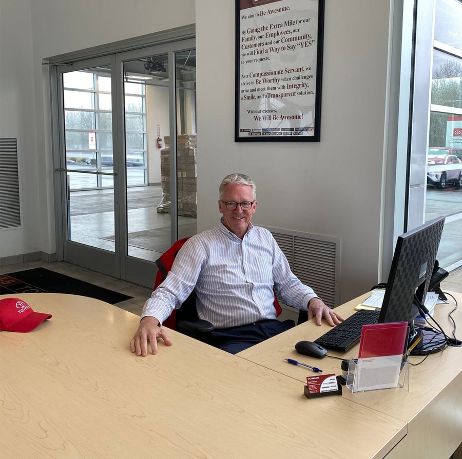 A man with glasses and gray hair sits smiling at a desk in an office with large windows. There’s a computer, notepad, and business cards on the desk. Cars and a parking lot are visible outside through the glass.