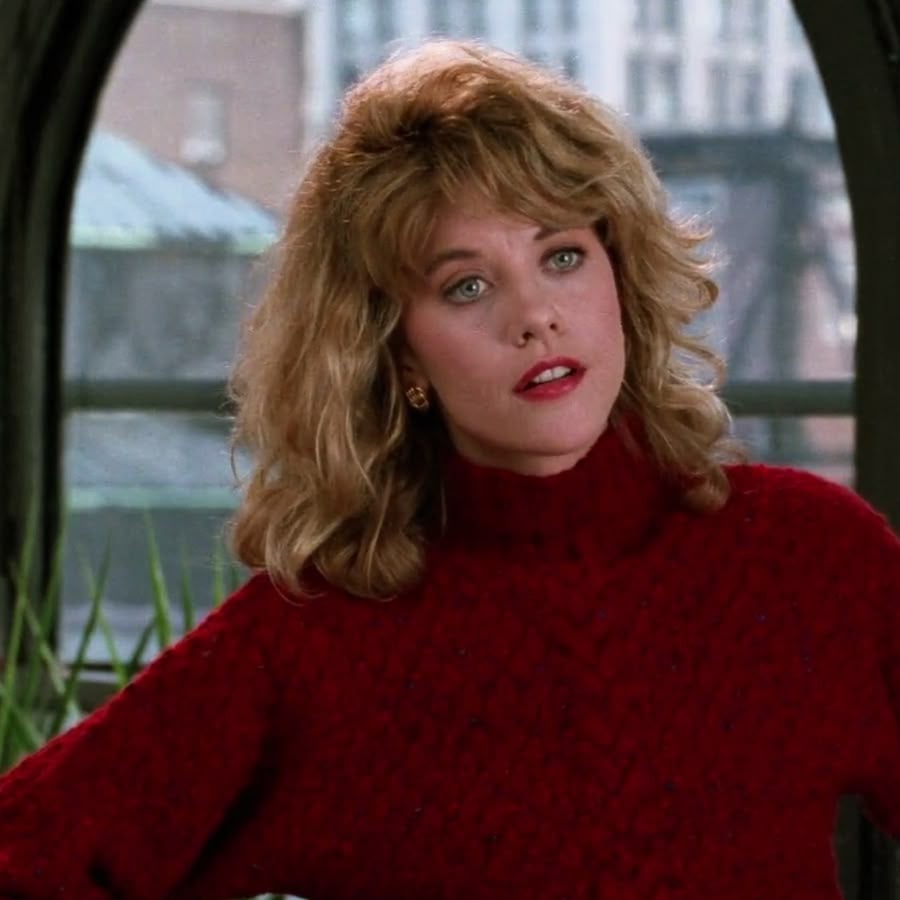 A woman with wavy blond hair and a red sweater stands indoors in front of a window, looking slightly to the side with a thoughtful expression. Urban buildings are visible outside the window.