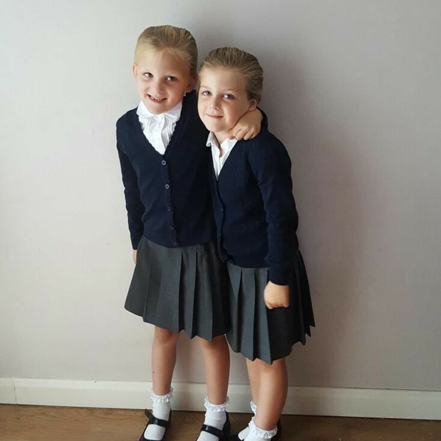 Two young girls stand side by side against a plain wall, wearing matching school uniforms with navy cardigans, white shirts, grey pleated skirts, white socks, and black shoes. One girl has her arm around the other.