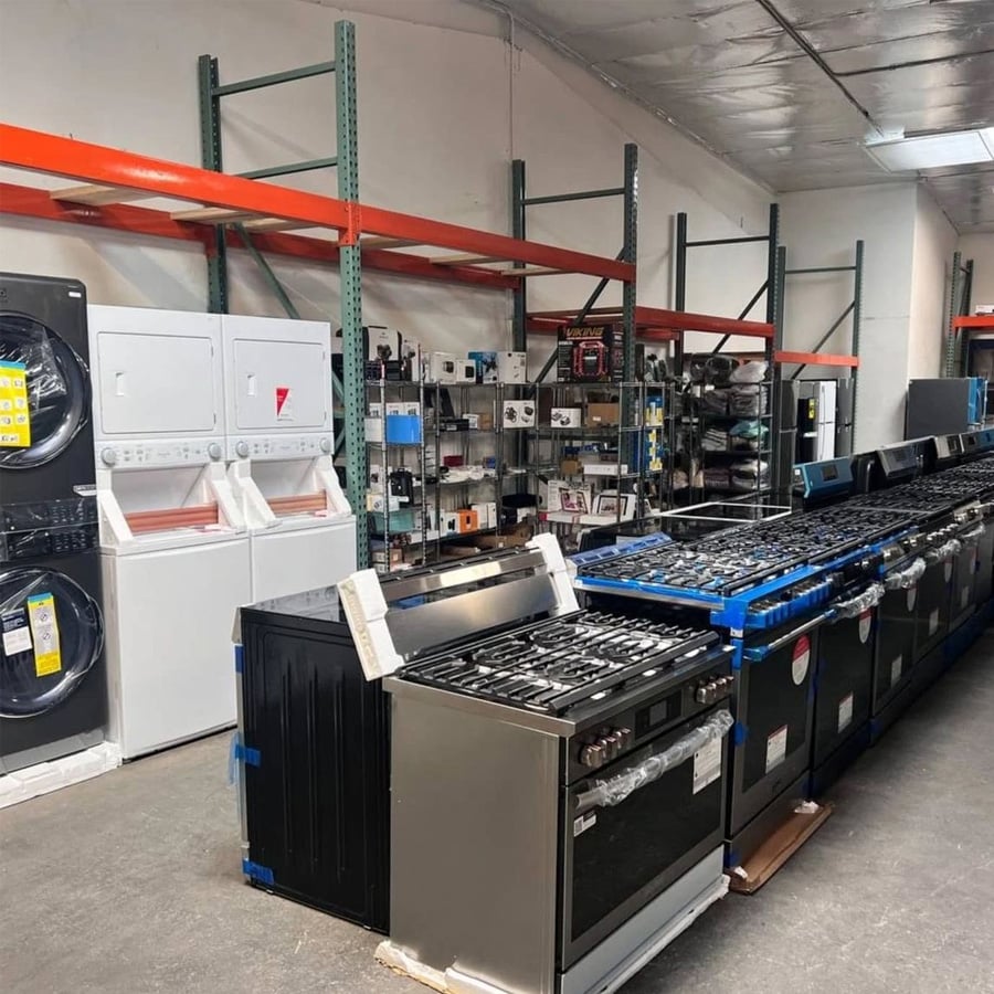 A warehouse displaying home appliances, including stacked washers and dryers, and rows of gas ranges and ovens. Metal shelves with various electronics and boxes line the back wall.