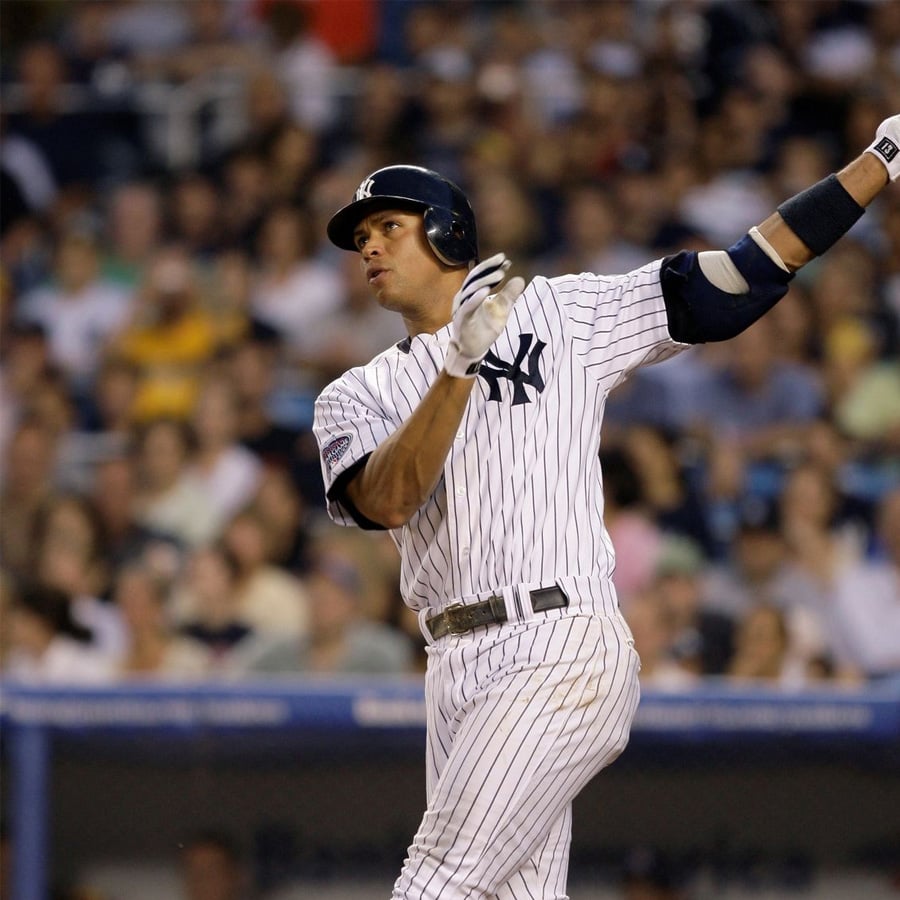 A baseball player in a New York Yankees uniform swings a bat during a game, with a crowd of spectators blurred in the background.